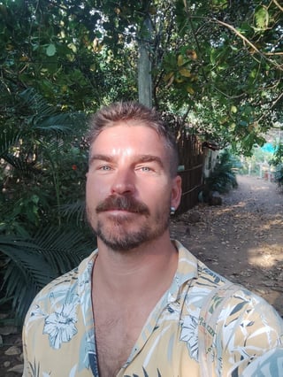 Man with dark hair and mustache wearing floral shirt, smiling outdoors among trees and greenery